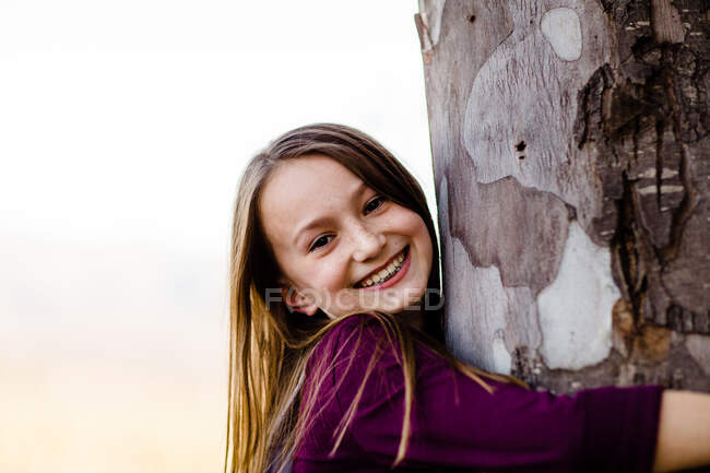 Giovane ragazza sorridente per fotocamera & abbraccio albero — Foto stock