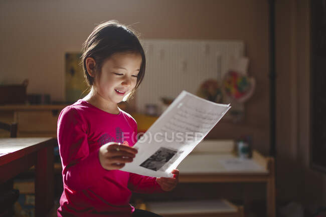 Un niño sonriente con una hermosa luz estudia un pedazo de papel, leyendo - foto de stock