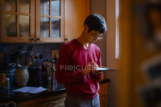 Un hombre está parado en una cocina junto a una ventana enviando mensajes de texto a un teléfono celular - foto de stock