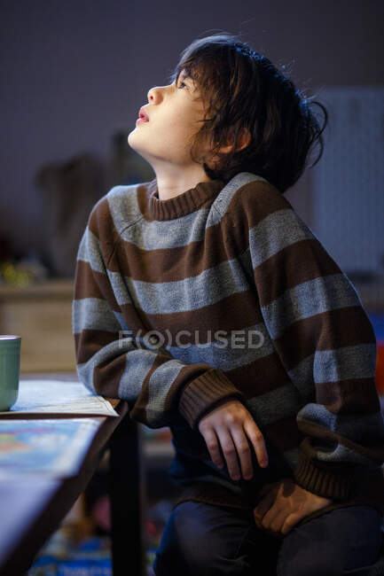 A beautiful boy in cozy sweater sits at table looking up into light — Stock Photo