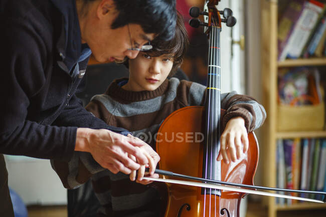 Un niño sosteniendo un violonchelo es enseñado por el padre a sostener correctamente un arco - foto de stock