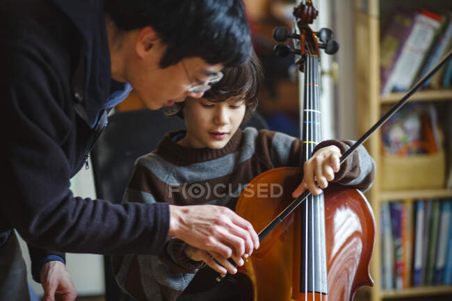 A father leans over child helping him learn to play a cello with a bow — Stock Photo