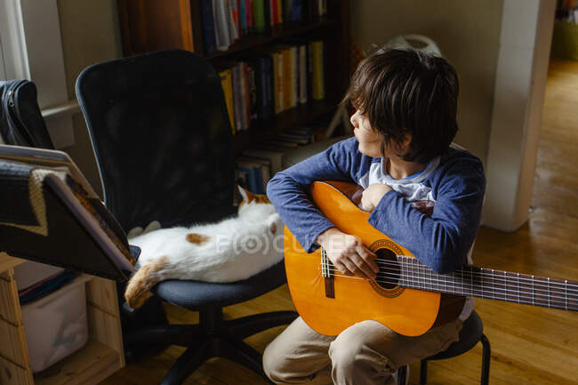 Un niño mira por una ventana sosteniendo una guitarra junto a un gato dormido - foto de stock