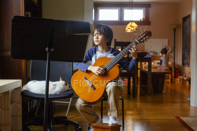 Хлопчик сидить тихо поруч з котом, граючи на гітарі, а сестра сидить позаду — стокове фото