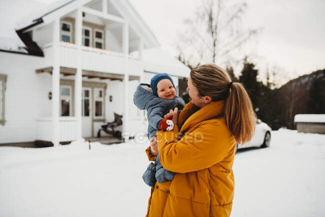 Mamá y adorable bebé sonriendo en frío día nevado fuera de la casa blanca - foto de stock
