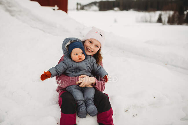 Sonriente hermana grande celebración bebé hermano fuera en la nieve en frío da - foto de stock