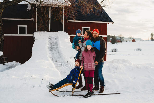 Linda família em pé junto ao celeiro vermelho no campo norueguês na neve — Fotografia de Stock