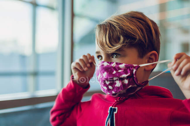 Мальчик в маске возле окна в аэропорту, чинит собственную маску — стоковое фото