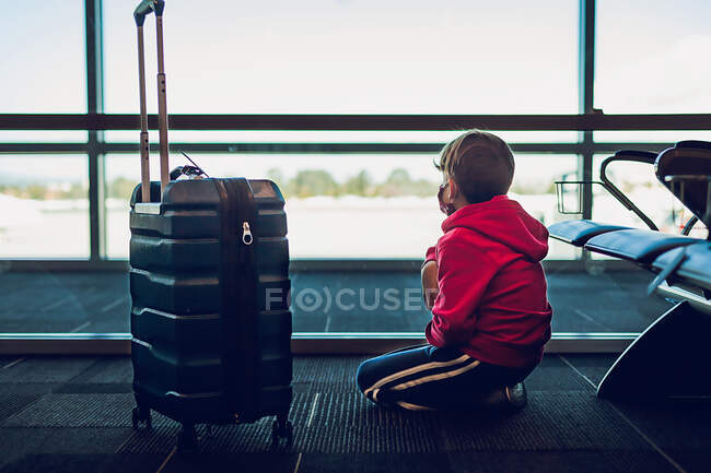 Niño al lado de la maleta mirando por la ventana en el aeropuerto - foto de stock