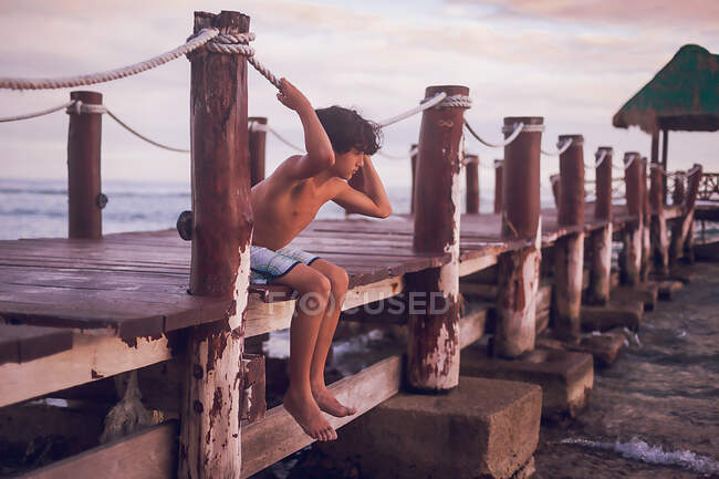 Pre adolescente niño sentado en un muelle de madera en una playa tropical - foto de stock