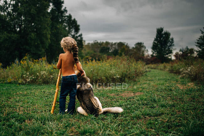 Menino vestindo chapéu de pele de casulo de pé com o cão em um campo — Fotografia de Stock