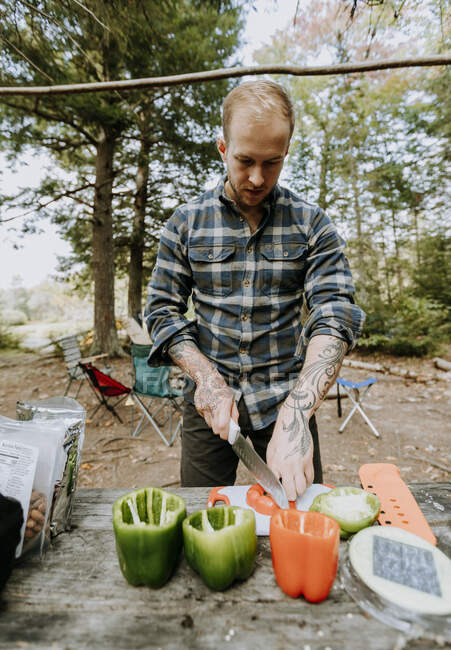 Man in flannel chops vegetables on table at campsite in woods, Maine — Stock Photo