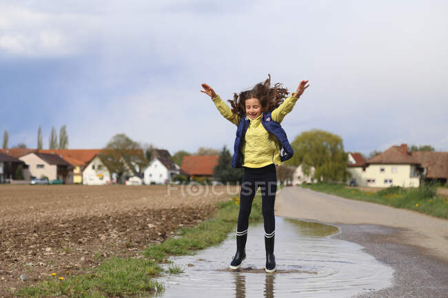 Una chica con los brazos levantados salta en un charco. - foto de stock