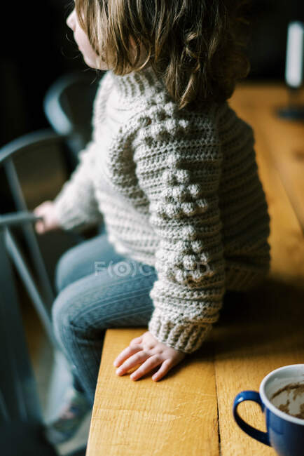 Niña sentada en la mesa en acogedor suéter de lana ganchillo casero - foto de stock