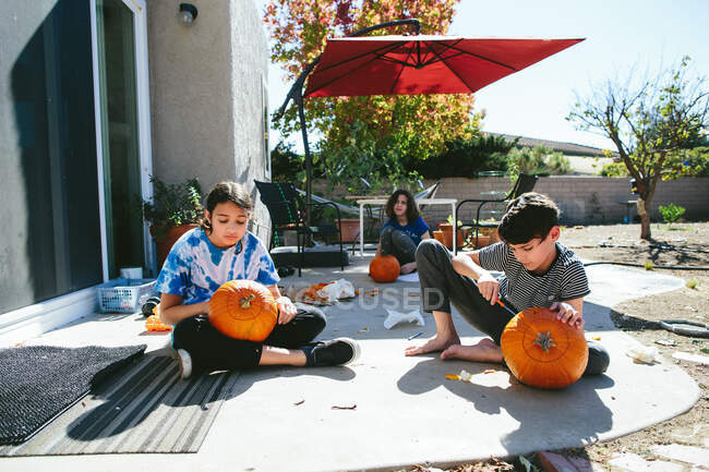 Tres niños se sientan afuera y tallan calabazas - foto de stock