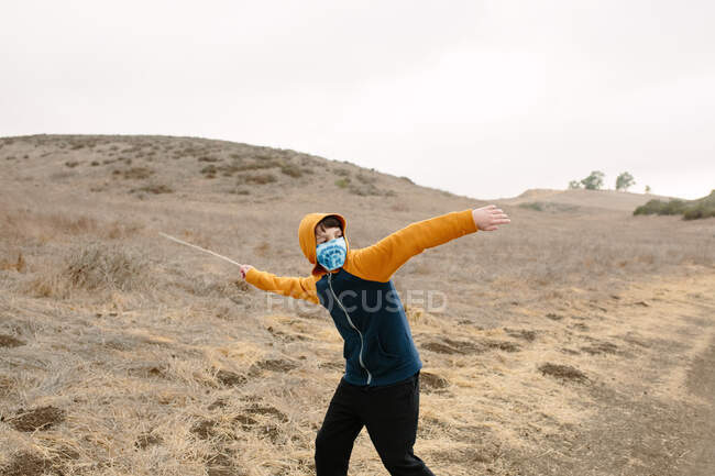 Niño en un sendero con una máscara de tela se prepara para lanzar un palo - foto de stock