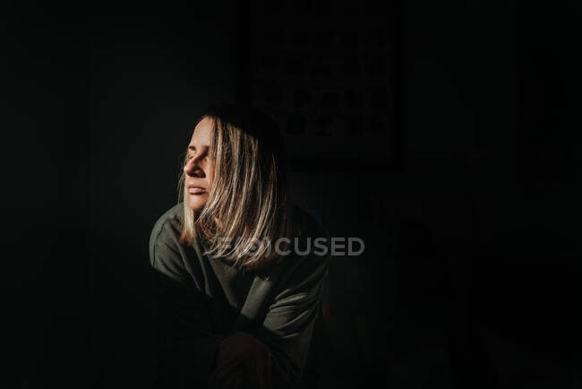 Mulher loira com fundo escuro se aquecendo na luz com os olhos fechados — Fotografia de Stock