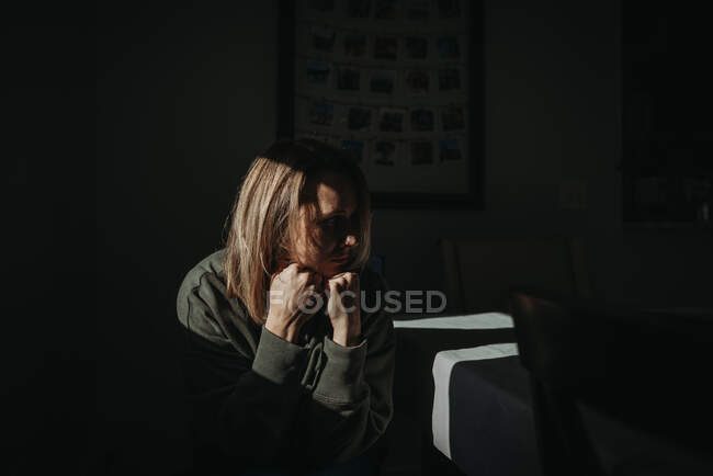 Adult woman sitting in sunlight with dark backdrop looking away sad — Stock Photo