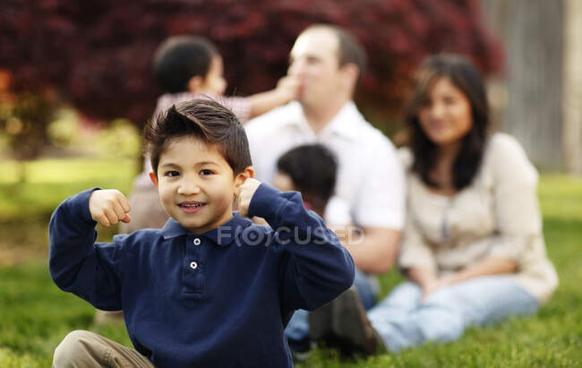 Menino flexionando enquanto sentado na frente da família na grama — Fotografia de Stock