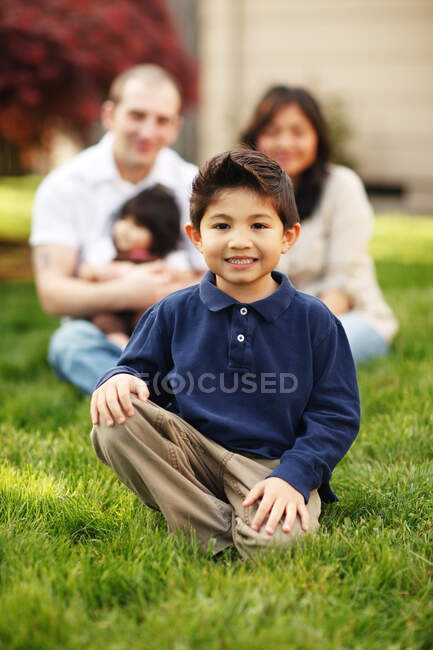 Niño sentado y sonriendo delante de la familia en la hierba - foto de stock