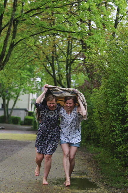 Due ragazze corrono scalze sotto un impermeabile e ridono. — Foto stock