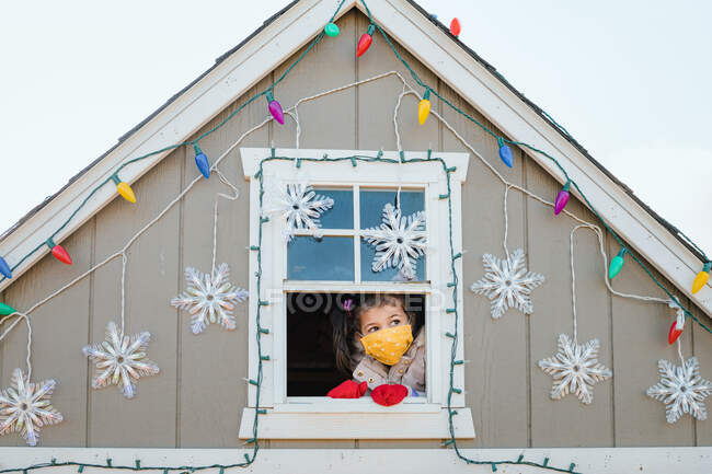 Chica joven en la máscara de la cara mirando por la ventana decoraciones de Navidad - foto de stock