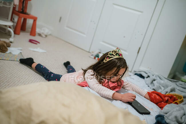 Chica joven con tiara jugando con el teléfono inteligente acostado - foto de stock