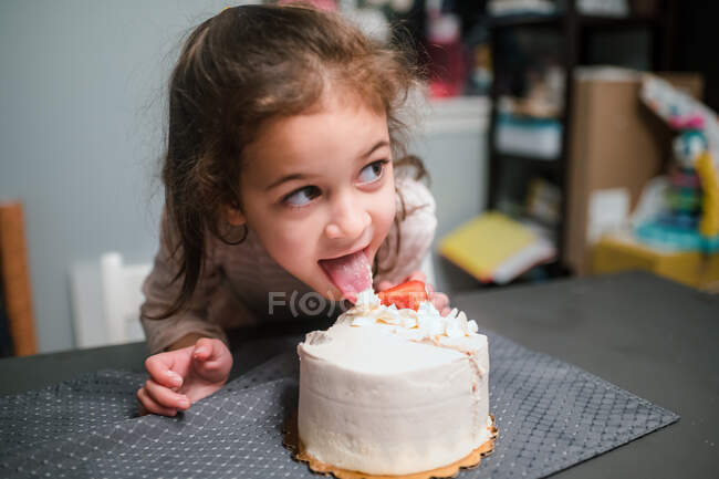 Edad preescolar chica lamiendo pastel de cumpleaños y mirando fuera de la cámara - foto de stock