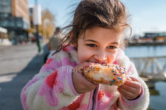 Chica joven comiendo donut con aspersiones fuera - foto de stock