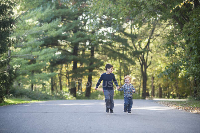 Irmãos alegremente de mãos dadas correndo por uma estrada treelined — Fotografia de Stock