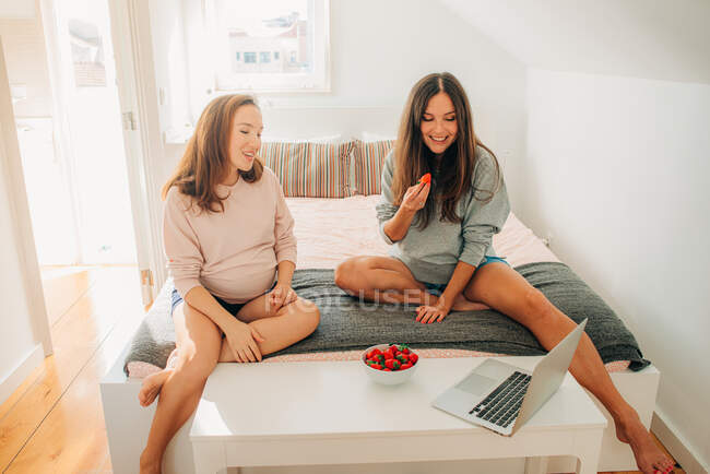 Two young pregnant women watching movie on laptop while sitting on bed and eating strawberries — Stock Photo