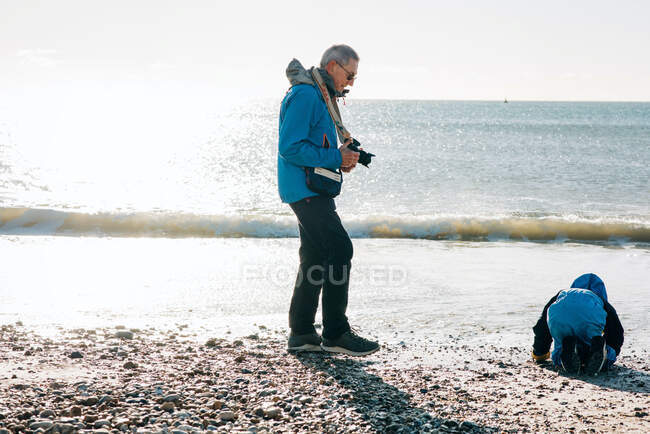 Abuelo tomando una foto de su nieto en un día soleado en la playa - foto de stock