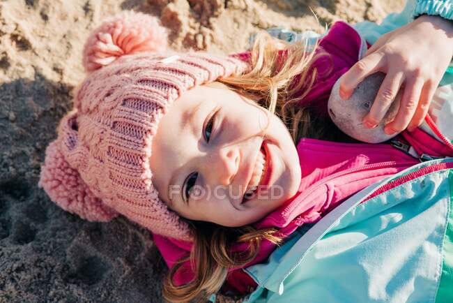 Junges Mädchen, das lächelnd mit ihren Steinen und Muscheln im Sand liegt — Stockfoto