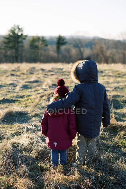 Двоє дітей стояли на вершині пагорба в зимовому одязі, щоб дивитися захід сонця — стокове фото