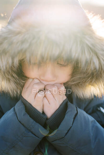 Niño con capucha soplando en las manos para luchar contra los dedos fríos de invierno - foto de stock