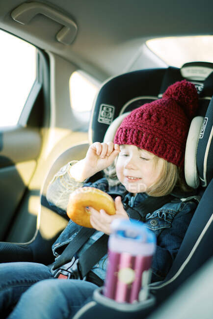 Petite fille heureuse avec chapeau rose jouit beignet dans le siège auto pendant le voyage — Photo de stock