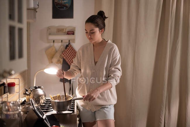 Молода жінка перемішує макарони в каструлі під час перегляду онлайн-рецепту вдома — стокове фото