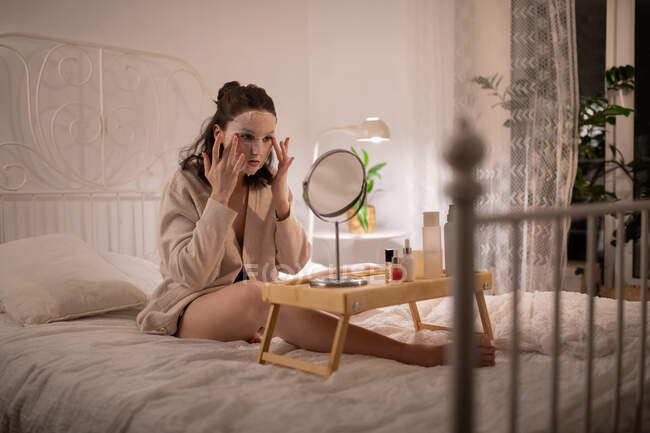 Giovane donna seduta sul letto e applicare maschera foglio durante la procedura di cura della pelle — Foto stock