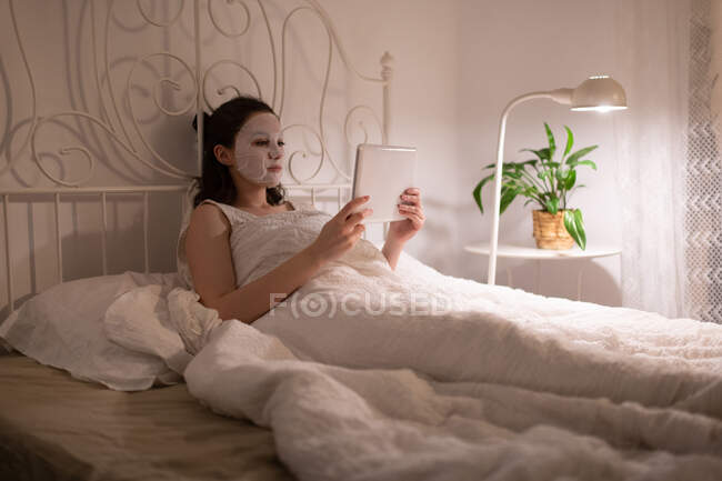Молода жінка з зволожуючою маскою спить на ліжку і дивиться фільм на планшеті — стокове фото
