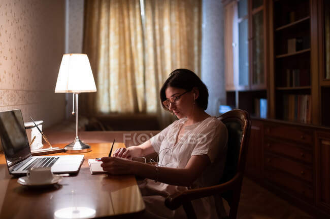 Jeune femme intelligente écoutant de la musique et écrivant dans un bloc-notes tout en étudiant à la maison — Photo de stock