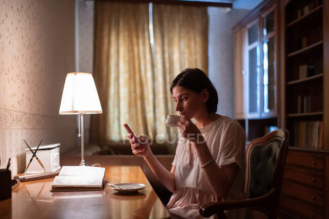 Молода жінка переглядає смартфон і сіє гарячий напій під час читання книги — стокове фото