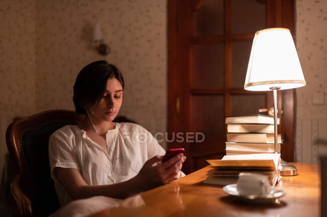 Young woman listening to music and using mobile phone while reading books at home — Stock Photo