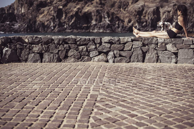Forte femme en bonne santé est assis avec petit chien au soleil chaud regardant l'océan — Photo de stock