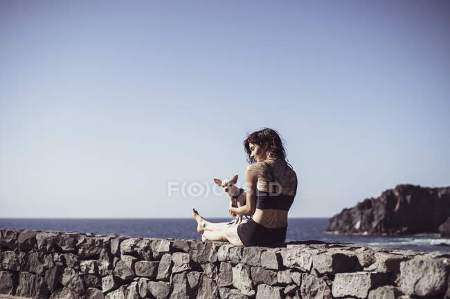 Mujer sana en forma con tatuajes se sienta junto al océano con en el sol con perro pequeño - foto de stock