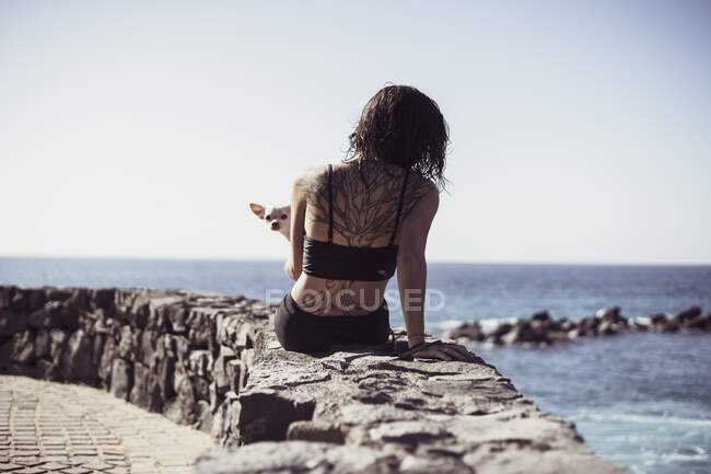Mujer en forma con tatuaje de árbol se sienta al borde del océano con lindo perro en el regazo - foto de stock