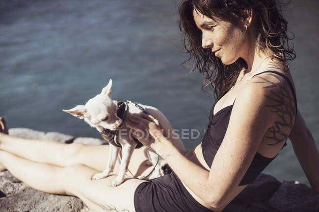 Healthy natural woman with freckles and tattoos sits with dog by ocean — Stock Photo