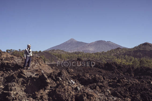Hiker with hoody stands on rocky volcano cliff looking out at mountain — Stock Photo