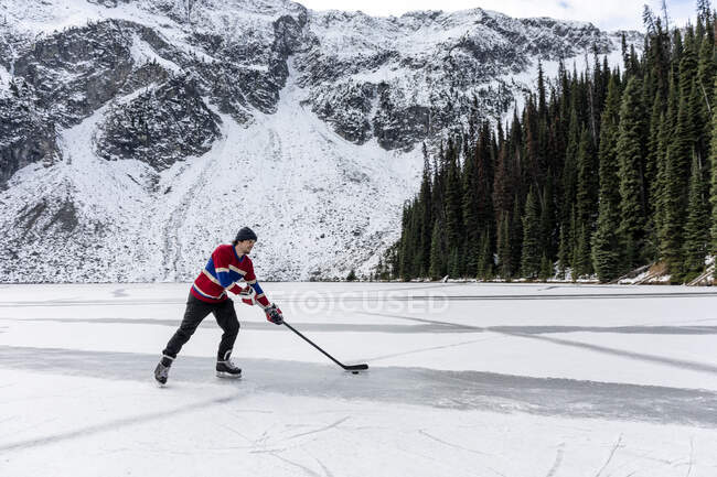Giovane giocatore di hockey cavalcando pattini da ghiaccio e manovra disco con bastone sul lago ghiacciato vicino montagna innevata e foresta di conifere in inverno in British Columbia, Canada — Foto stock