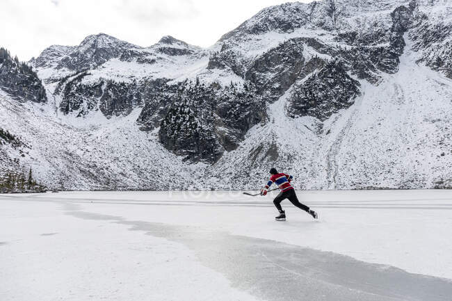 Человек играет в хоккей возле заснеженных гор на замерзшем озере — стоковое фото