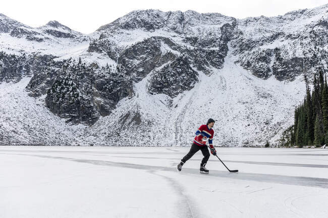 Молодой человек играет в хоккей на озере возле заснеженной горы — стоковое фото